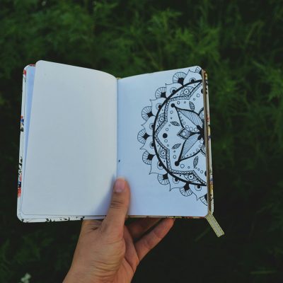 A hand holding an open journal with a detailed mandala drawing outdoors.