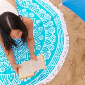 A woman lies on a colorful beach blanket reading a book on sandy shore under a clear sky.